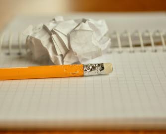 Orange penna med ett suddgummi på toppen som ligger på ett randigt kollegieblock. Bredvid ligger en ihopskrynklad pappersboll.